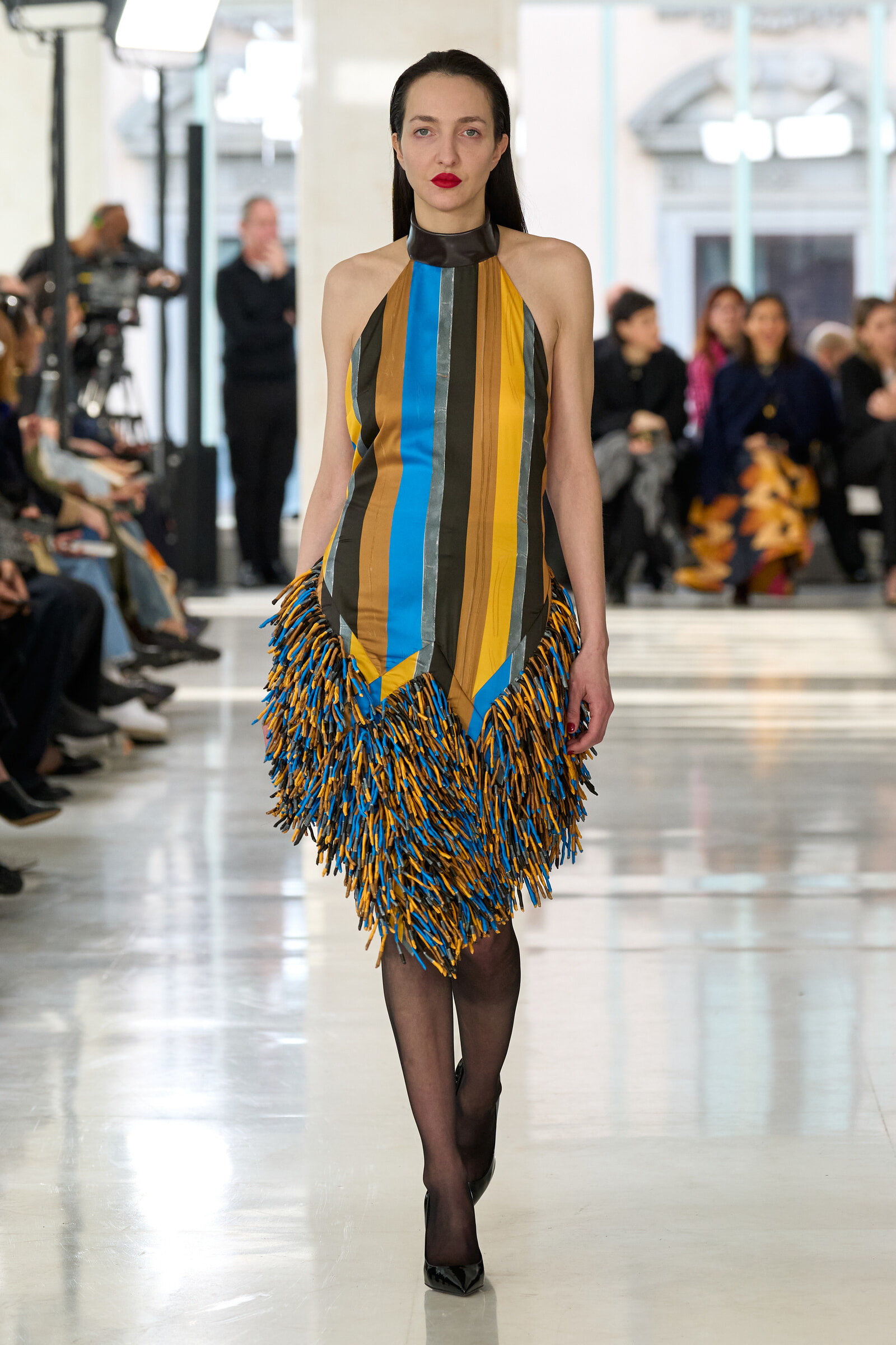 Model walking the runway in a sleeveless vertical-striped dress with blue, yellow, brown, and gray panels and bold fringe; audience in background.