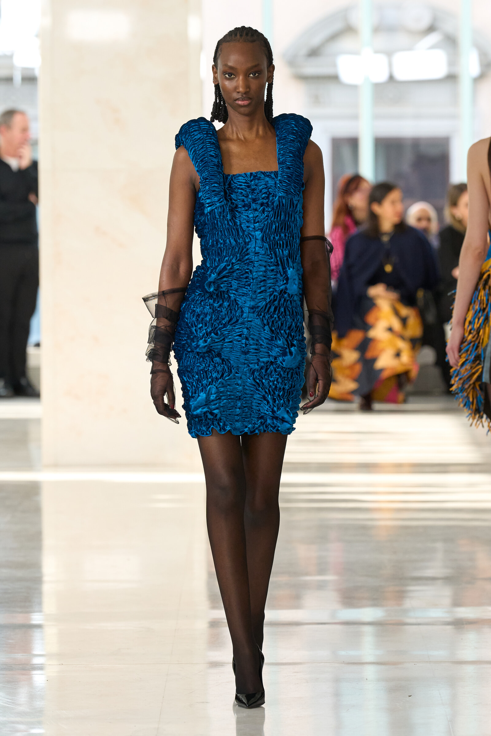 Model walks a fashion runway in a bright blue textured ruched dress and sheer black gloves.