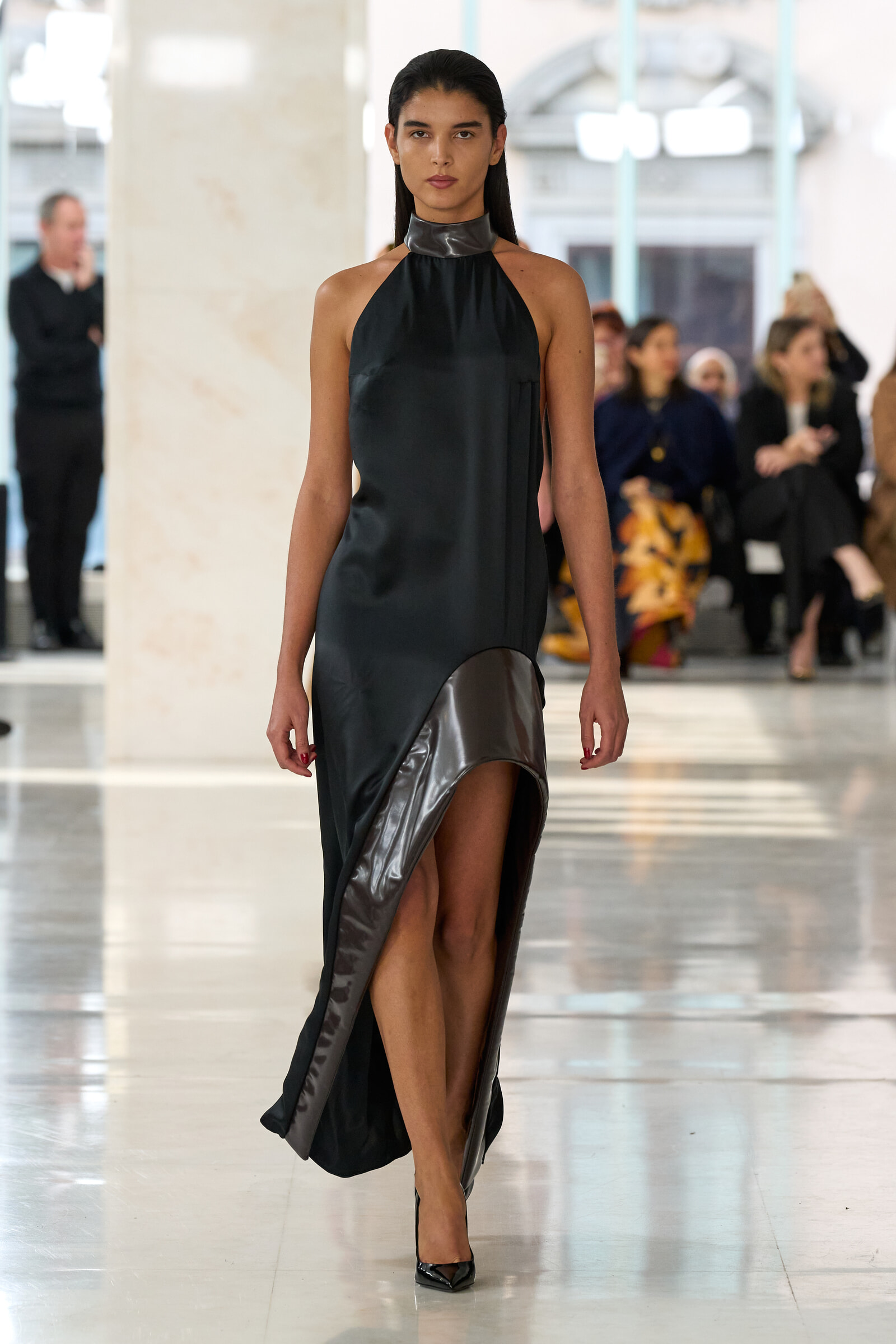 Model walks the runway in a black halter dress with metallic accents, as blurred fashion-show audience watches in the background.
