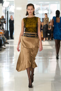 Model on a runway wearing a mustard sleeveless top with black thread embellishments and a beige satin skirt with stacked belts, walking forward.