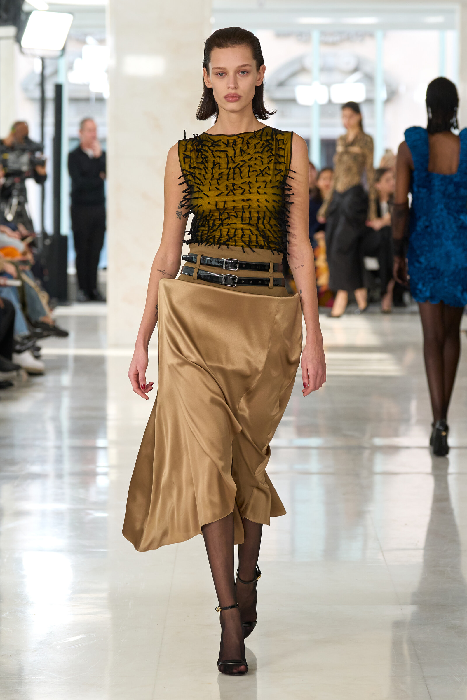 Model on a runway wearing a mustard sleeveless top with black thread embellishments and a beige satin skirt with stacked belts, walking forward.