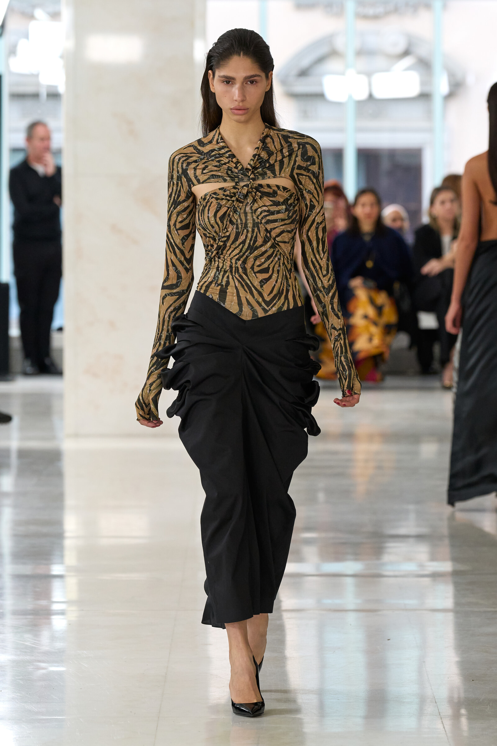 Model walks a runway in a zebra-print sheer top with cutouts and a black draped skirt, black heels.