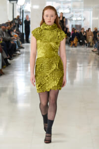 Model walking a green crushed-velvet dress down a runway during a fashion show, audience seated on both sides in background.