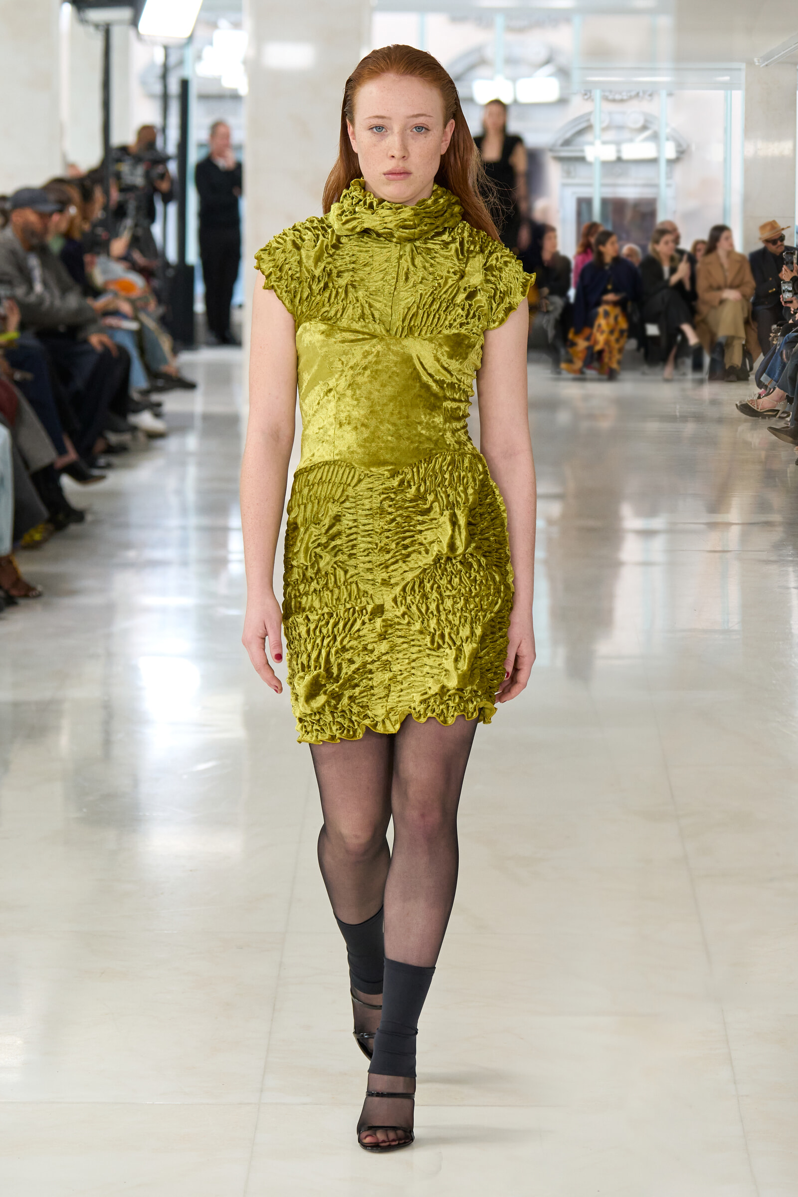 Model walking a green crushed-velvet dress down a runway during a fashion show, audience seated on both sides in background.