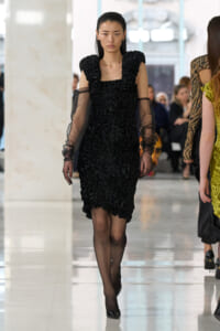 Model walks a fashion runway in a black textured dress with voluminous shoulders and sheer long sleeves in a bright indoor venue.