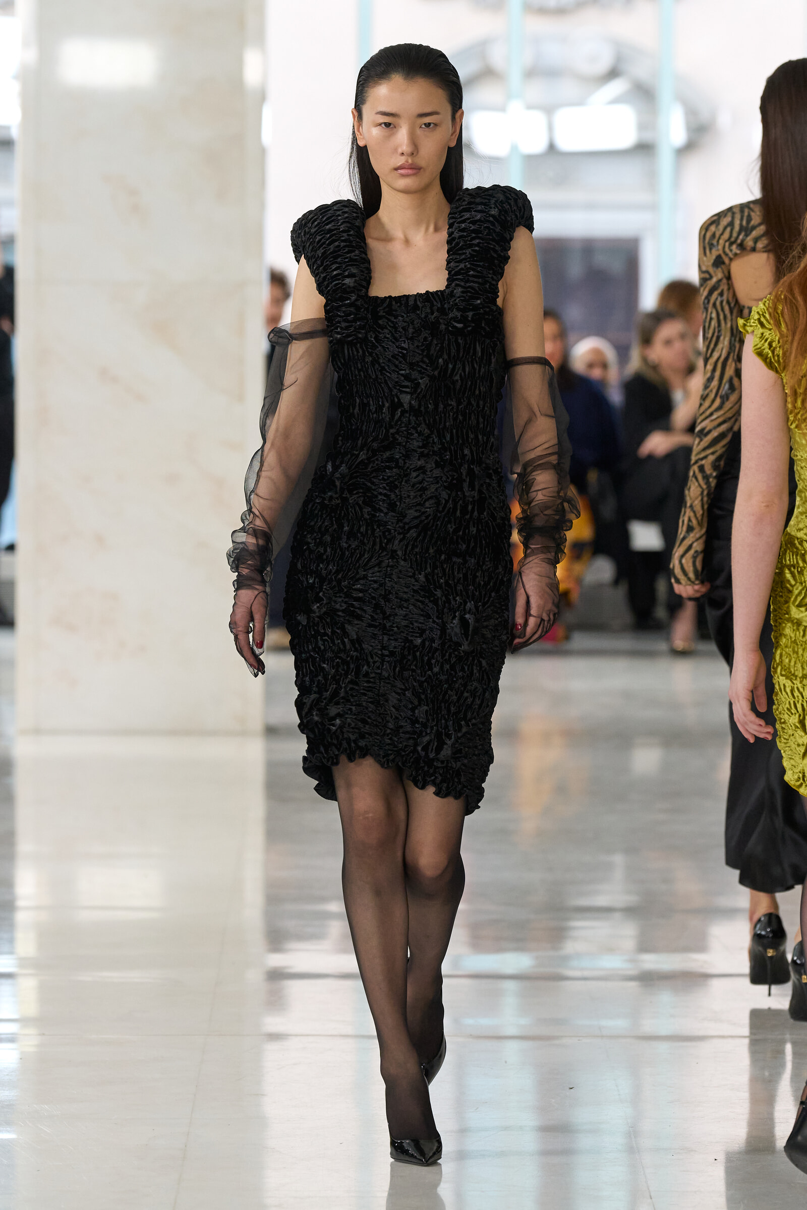 Model walks a fashion runway in a black textured dress with voluminous shoulders and sheer long sleeves in a bright indoor venue.