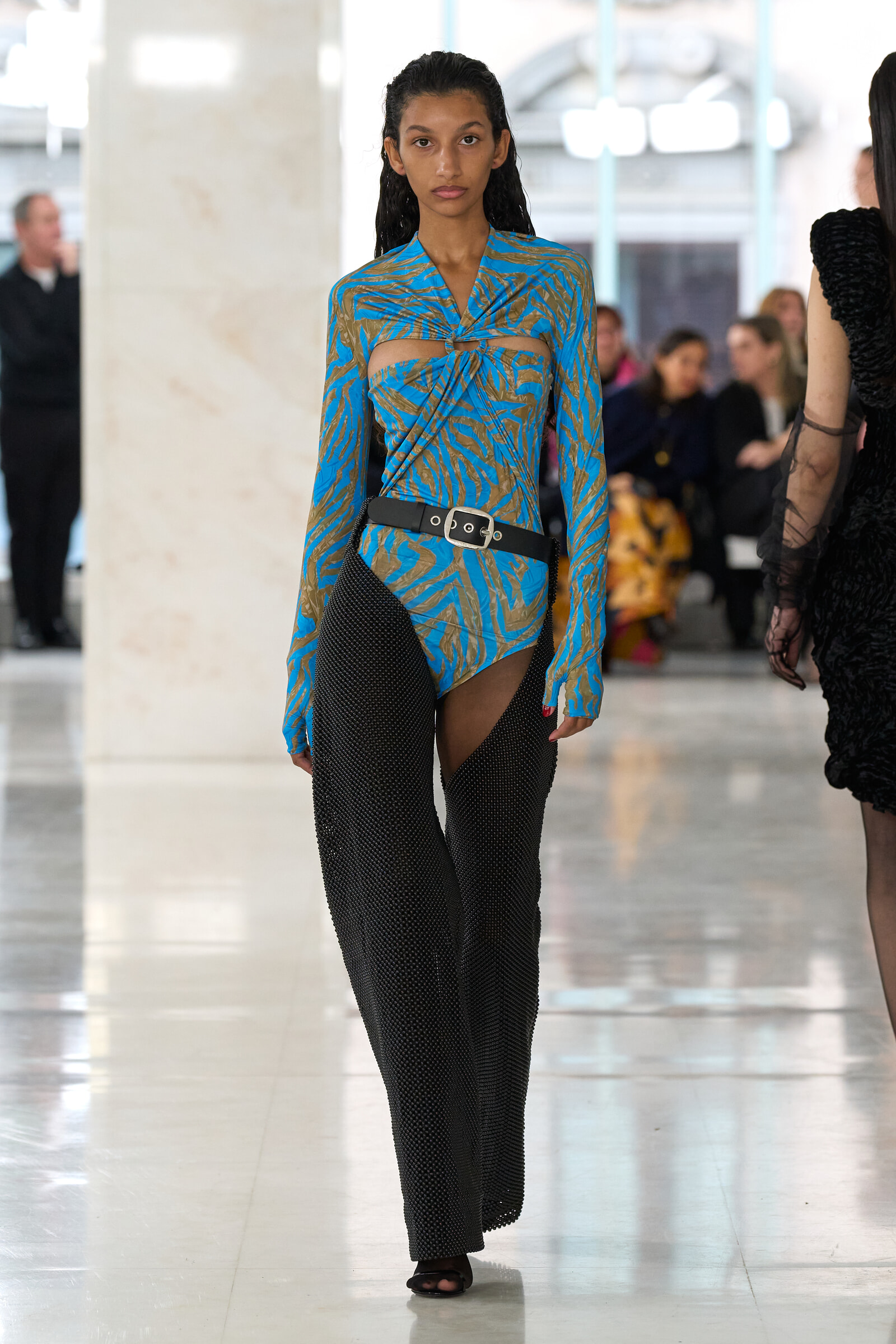 Female model walking a runway in a blue tiger-print top with cutouts and black mesh pants, cinched with a wide belt.