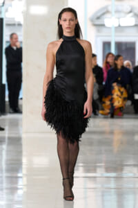 Female model in a black satin halter-neck dress with fringe walks down a runway, blurred audience in the background.
