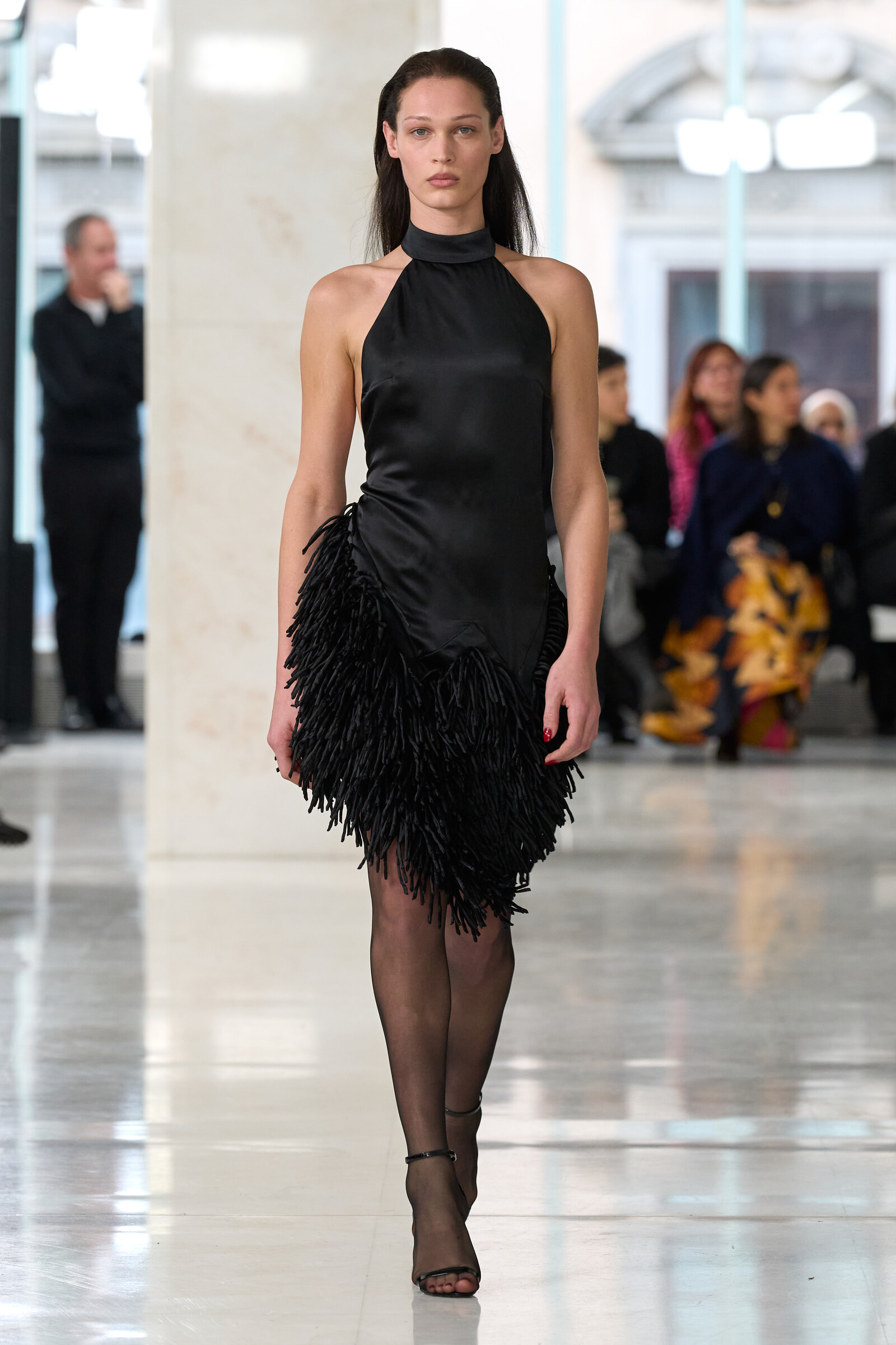 Female model in a black satin halter-neck dress with fringe walks down a runway, blurred audience in the background.