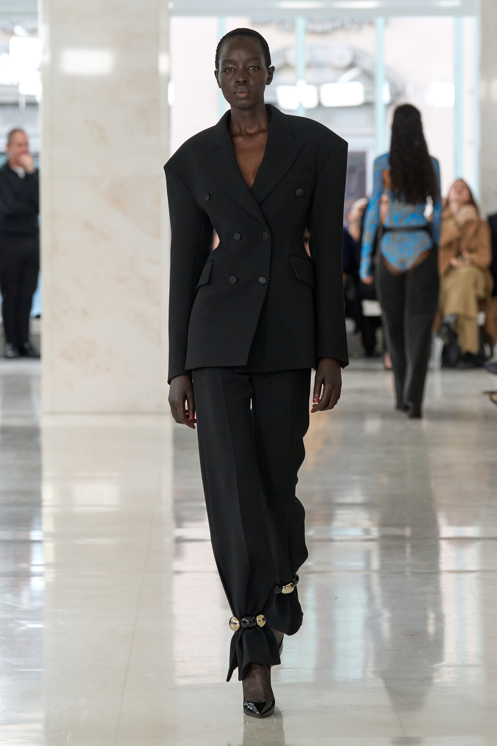 Runway model wearing a structured oversized black double-breasted suit with wide shoulders, walking in a bright indoor venue.