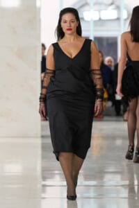 Runway model walking toward camera in a black V-neck dress with sheer sleeves and red lipstick.