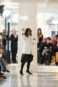 Model in a white blazer and black skirt waves from behind a marble pillar on a runway as photographers and audience applaud.