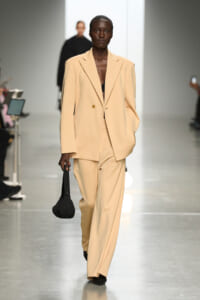 Model walks a beige oversized blazer and matching wide-leg suit on a runway, carrying a black bag with her left hand.