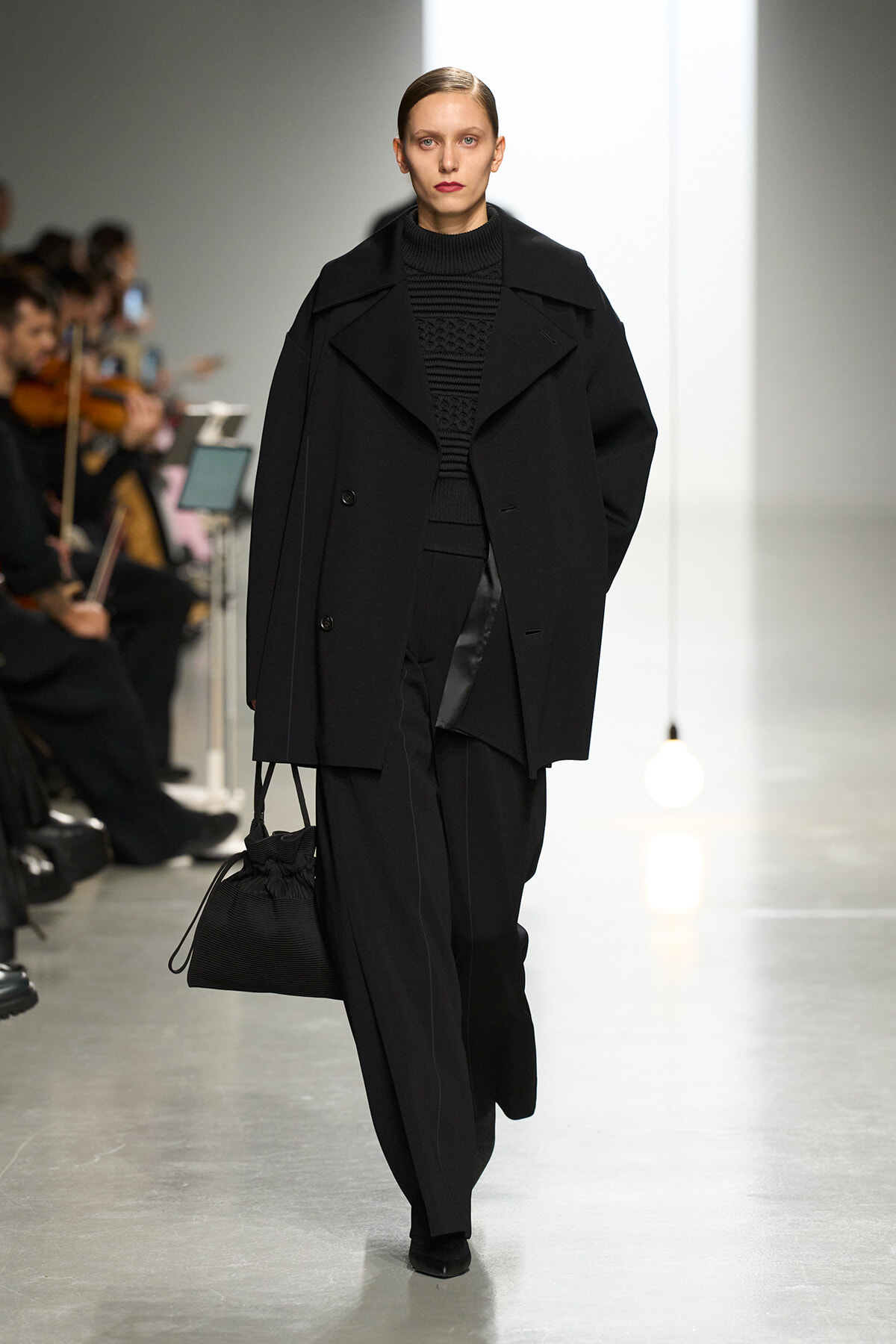 Model on a fashion runway wearing a black oversized coat and black outfit, carrying a black bag with a blurred audience in the background.
