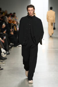 Male model walks a fashion runway in an all-black layered outfit with oversized coat and glasses; audience lines the catwalk left.