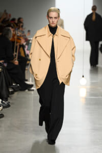 Model walks the runway in a camel oversized cape coat over a black turtleneck and wide-leg black pants; audience blurred in the background.