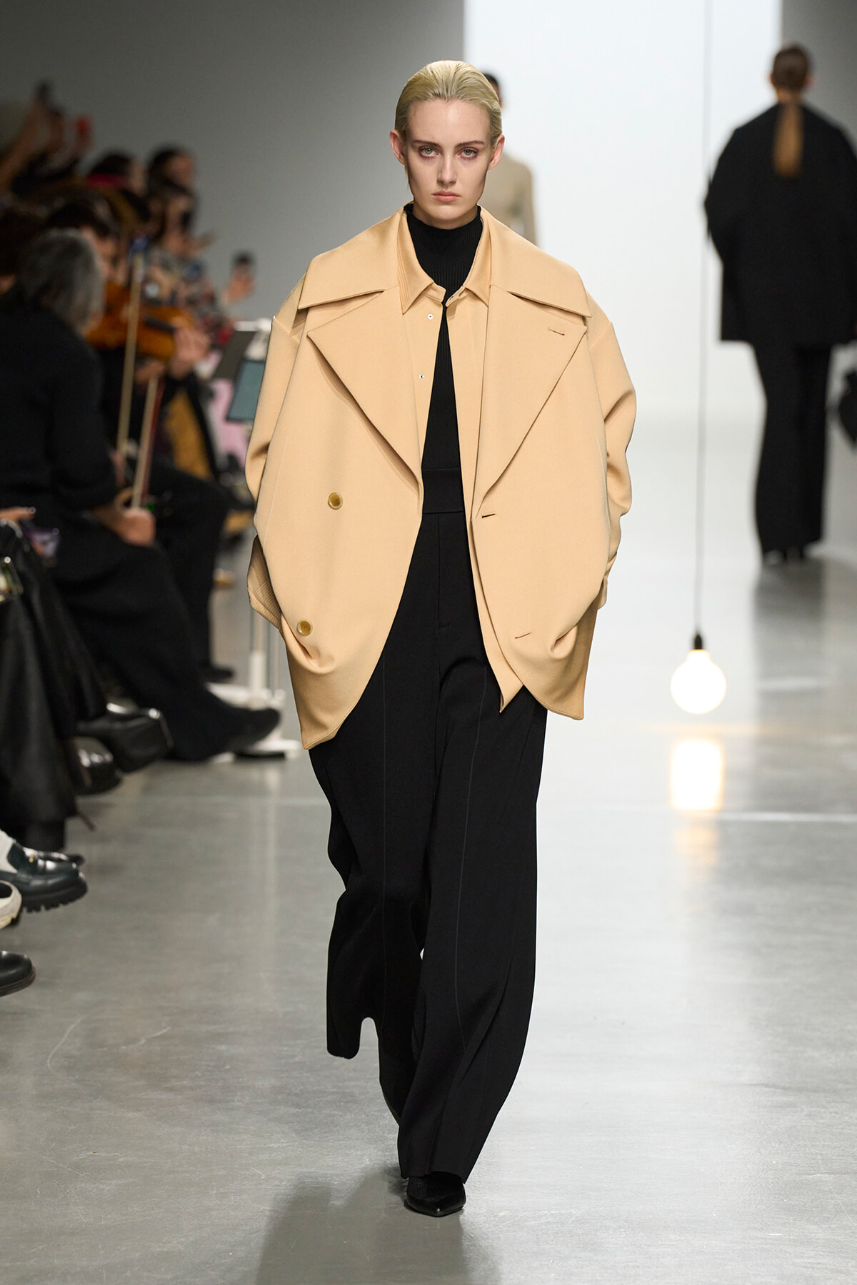 Model walks the runway in a camel oversized cape coat over a black turtleneck and wide-leg black pants; audience blurred in the background.