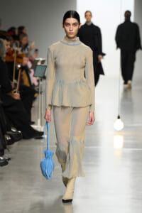 Model walks a fashion runway in a pale beige ribbed knit peplum top with blue accents and matching wide-leg pants, carrying a light blue drawstring bag.