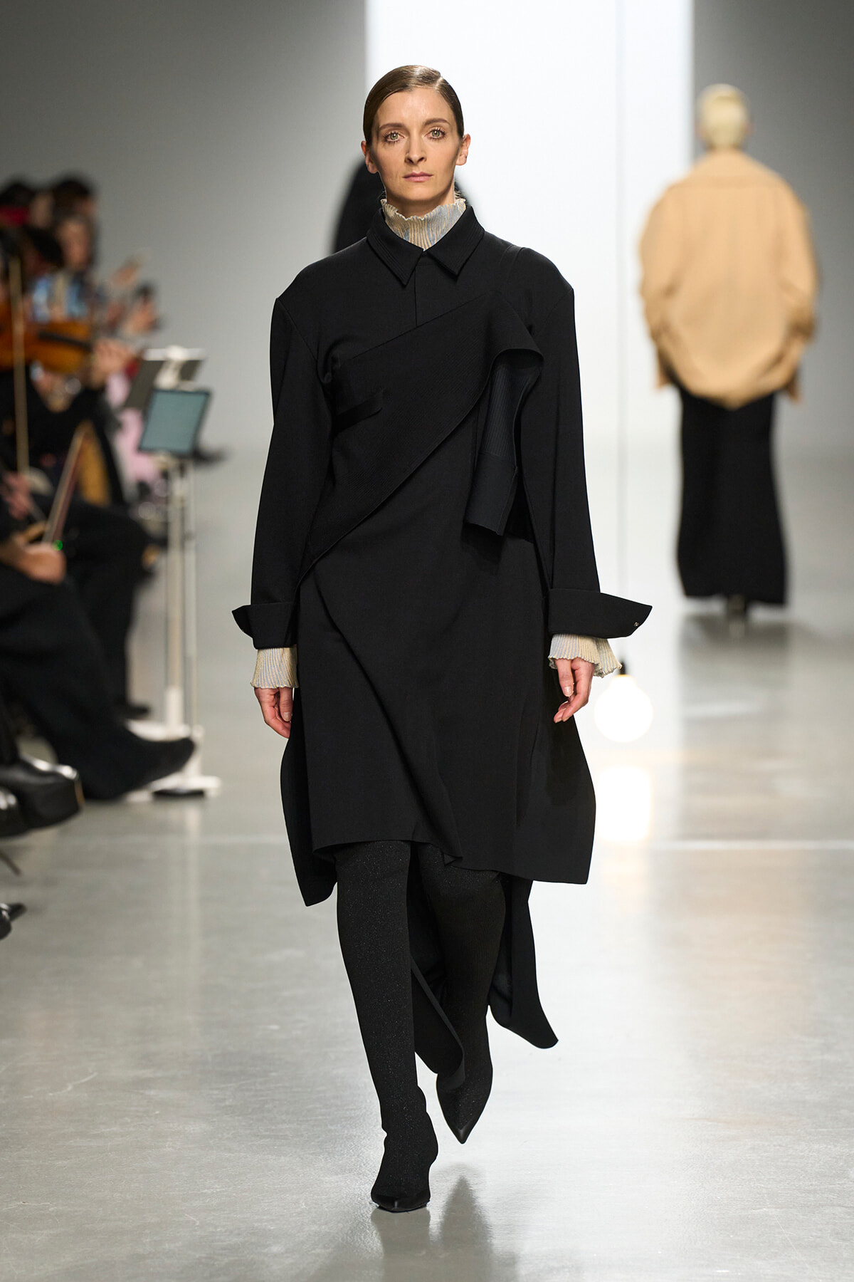 Model walks the runway in a black layered wrap coat with diagonal panels, beige knit cuffs peeking out, audience on the left.