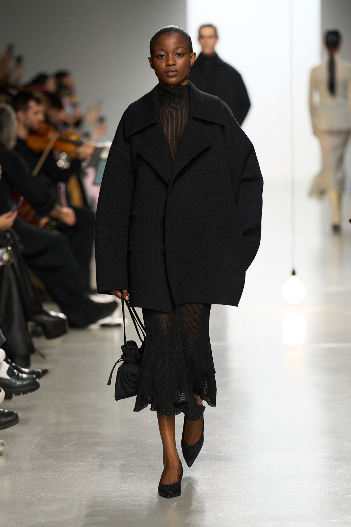 Model walks the runway in an oversized black coat over a sheer black top and black pleated skirt, with pointed heels.