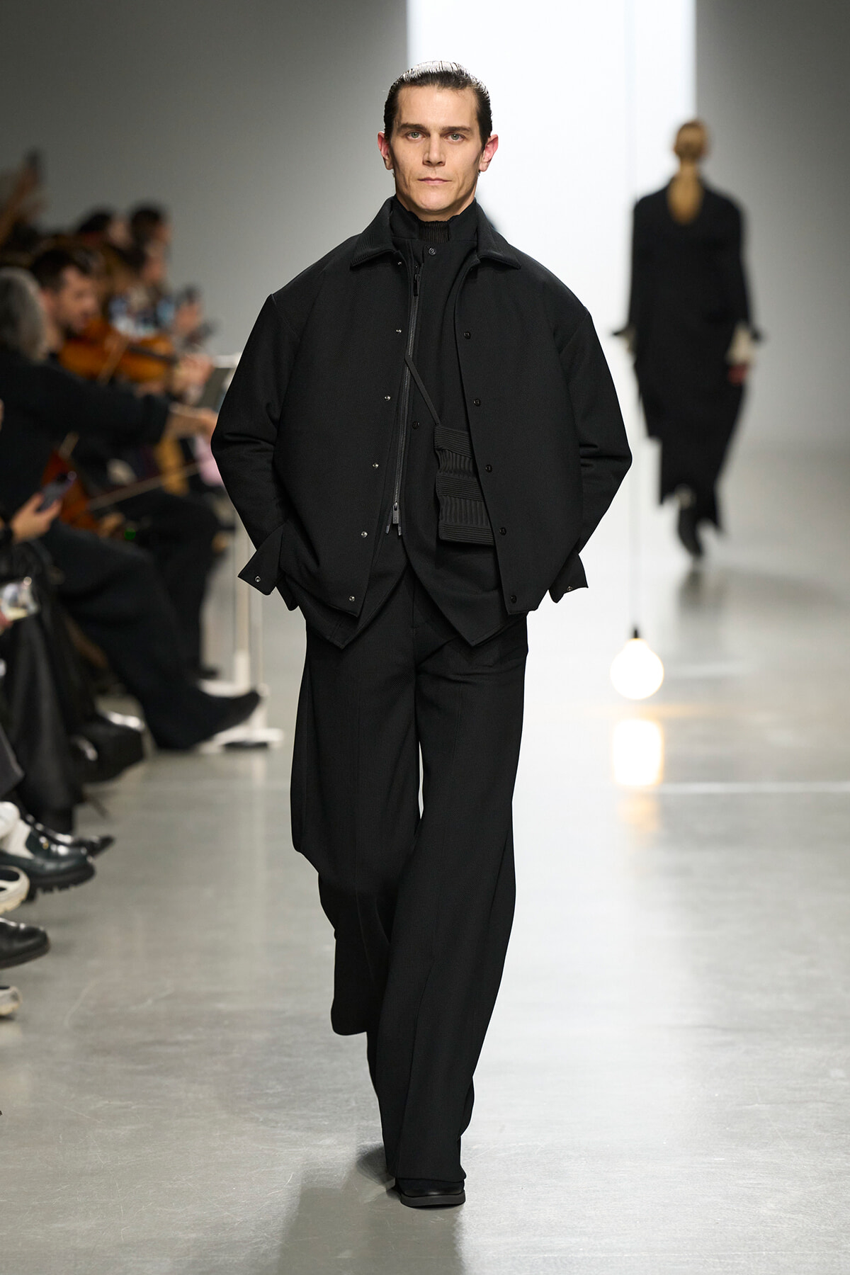 Male model walking a runway in an all-black layered outfit with a long coat and wide-leg pants; audience visible on the left.