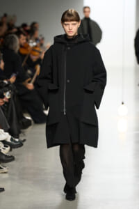 Model on a runway wearing an oversized black coat with a front zipper and large pockets, hands in pockets, audience blurred in the background.