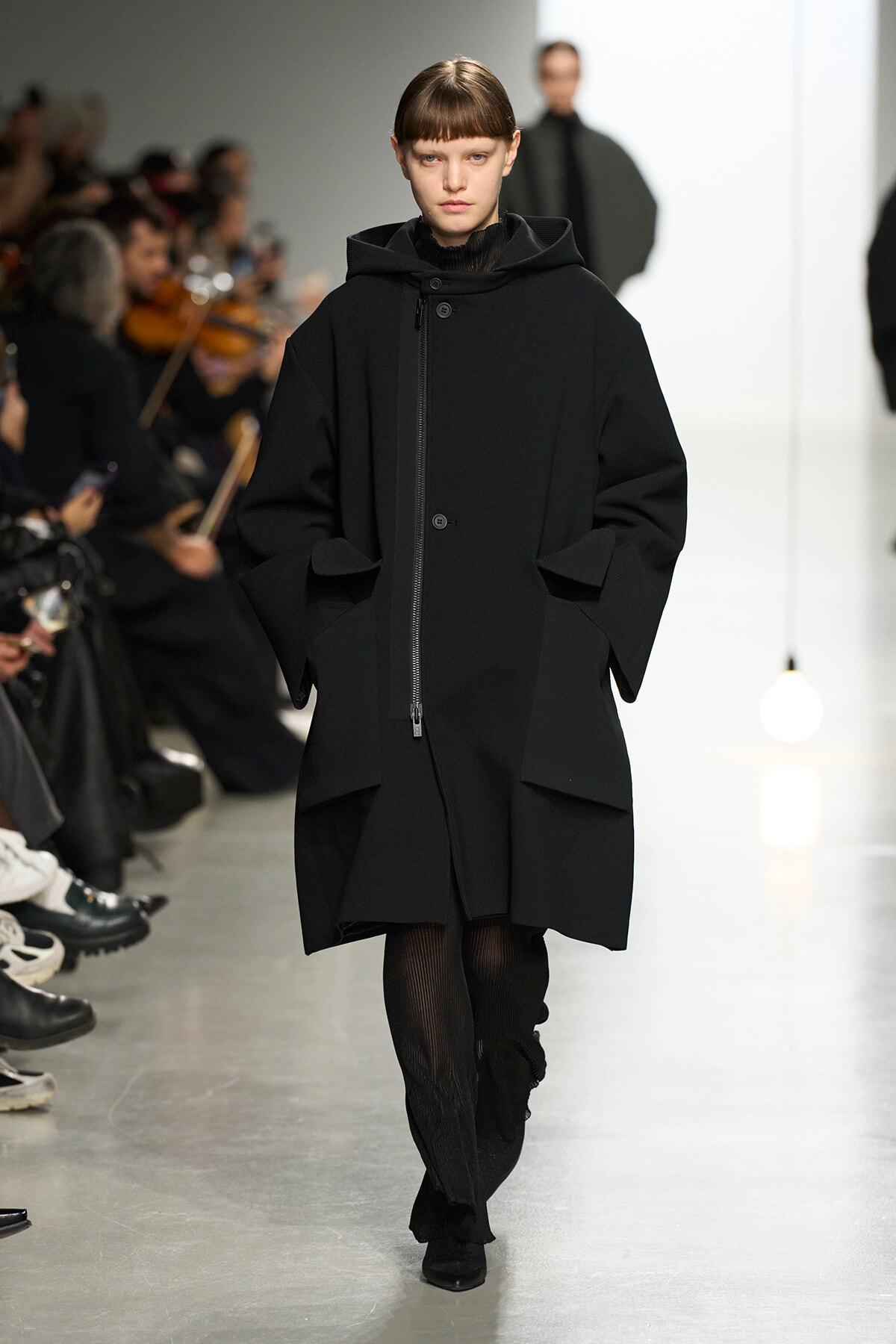 Model on a runway wearing an oversized black coat with a front zipper and large pockets, hands in pockets, audience blurred in the background.
