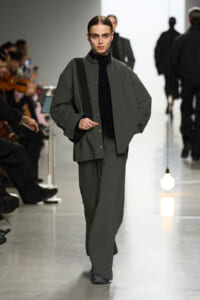 Model walks the runway in an oversized gray jacket and matching wide-leg pants with a black turtleneck as a crossbody strap rests on her shoulder, minimalist styling.