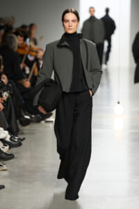 Runway model wearing a gray zip-front jacket over a black turtleneck and wide black pants, carrying a black bag as audience watches.