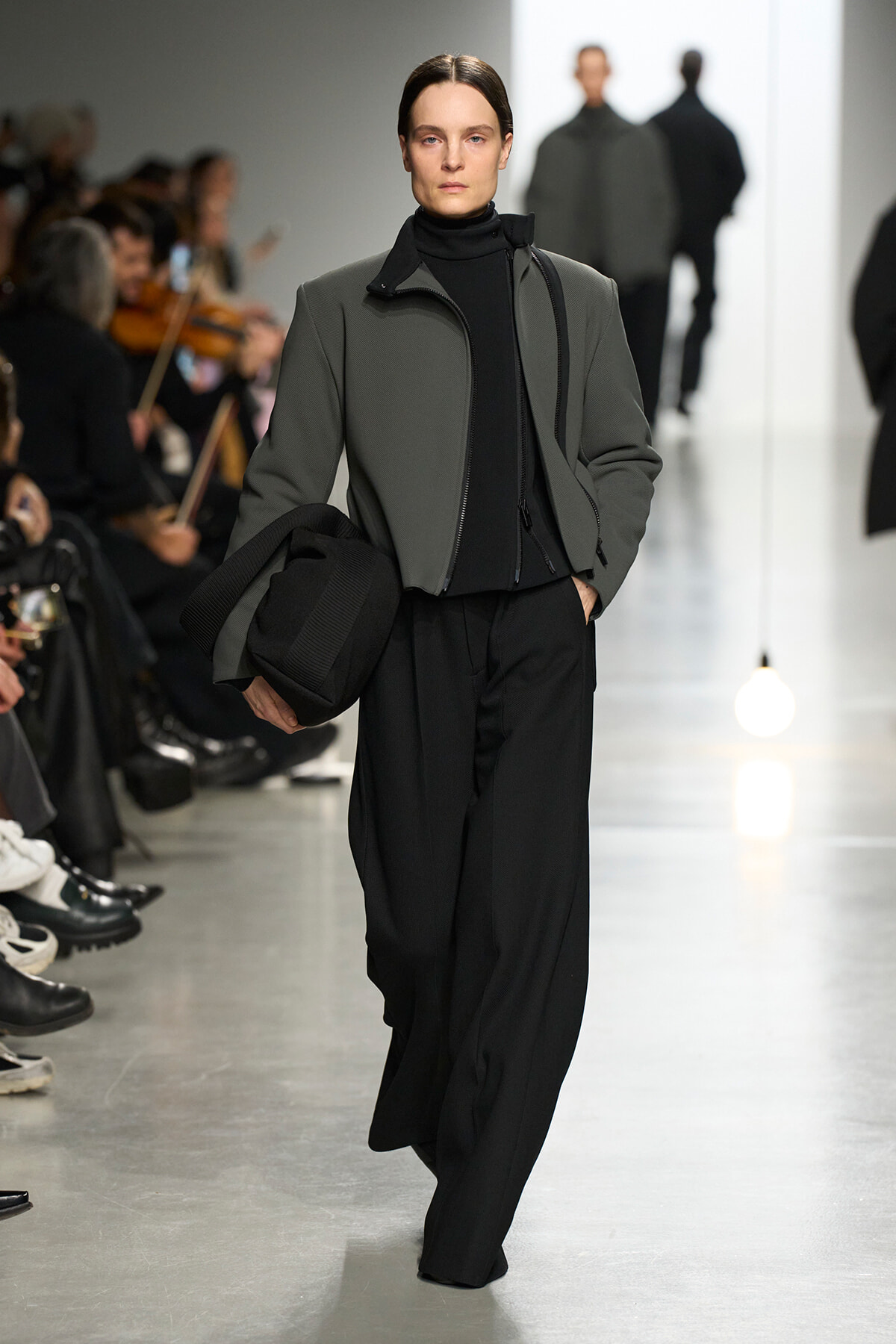 Runway model wearing a gray zip-front jacket over a black turtleneck and wide black pants, carrying a black bag as audience watches.