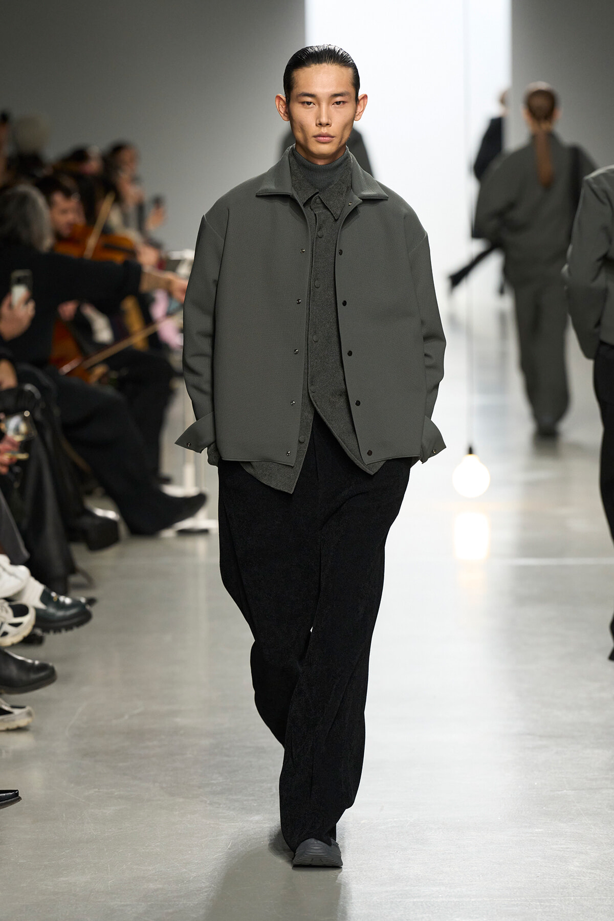 Model walking the runway in a gray layered jacket over a dark turtleneck and wide-leg pants, audience watching on either side.