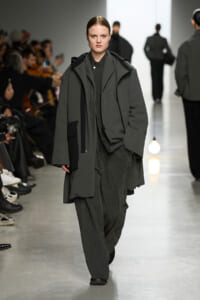 Model walks a dark, oversized gray-green coat down a fashion runway with audience on either side.