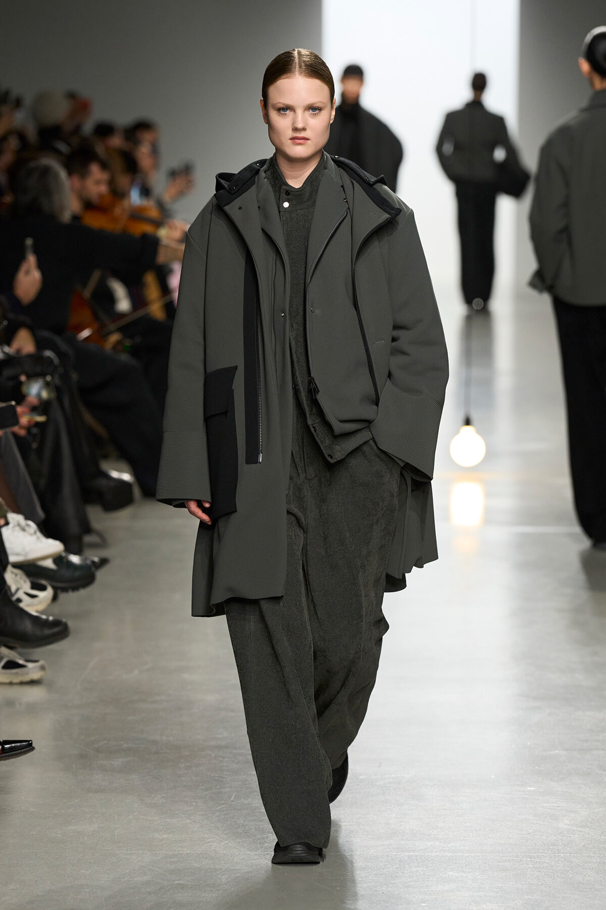 Model walks a dark, oversized gray-green coat down a fashion runway with audience on either side.