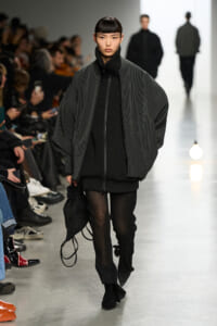 Model walking a fashion runway in an oversized striped gray jacket over a black knit outfit, carrying a black bag in one hand.