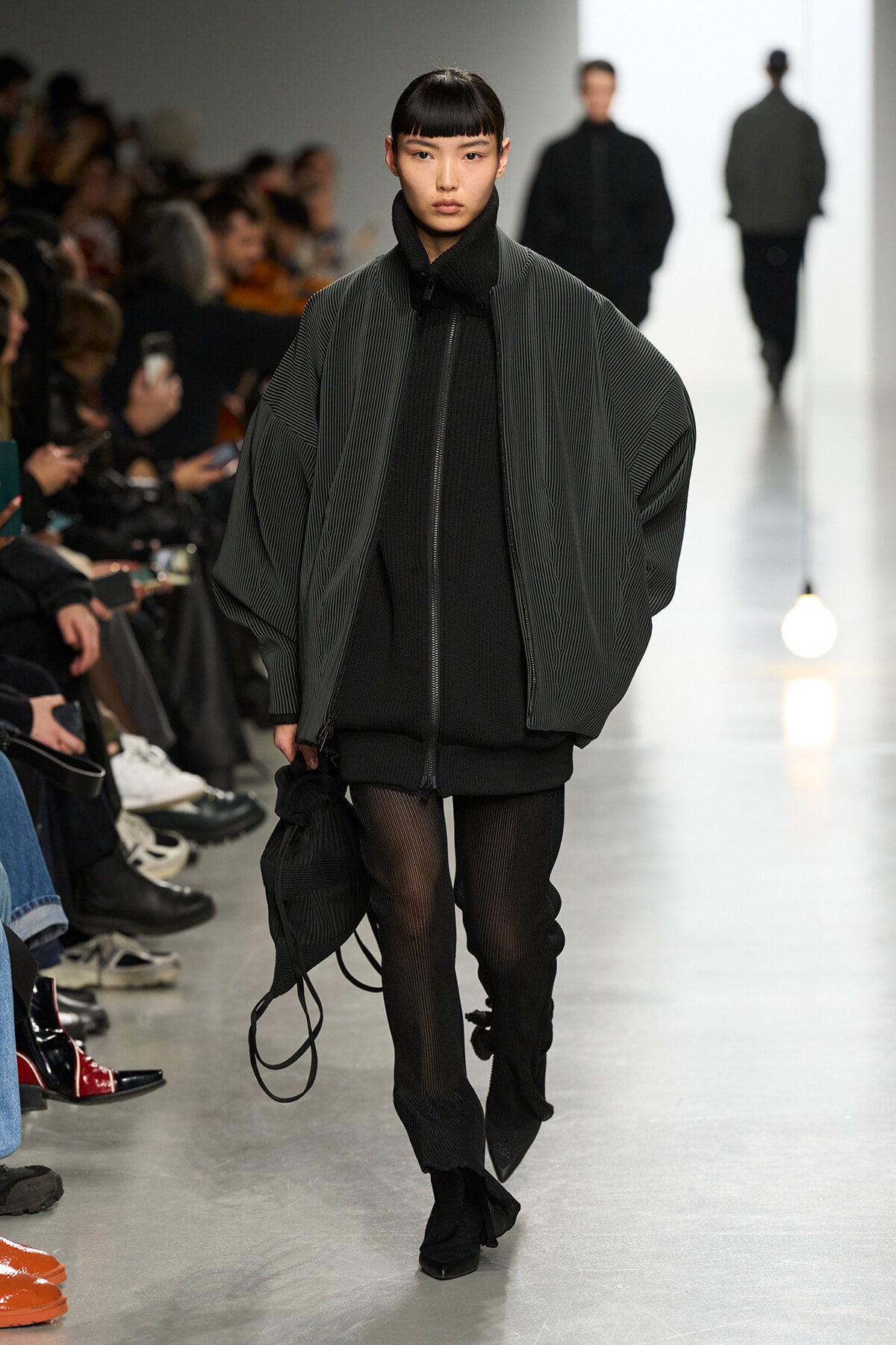 Model walking a fashion runway in an oversized striped gray jacket over a black knit outfit, carrying a black bag in one hand.