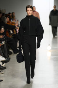 Model walking a runway in an all-black ribbed zip-front top with oversized sleeves and matching pants, holding a black bag.