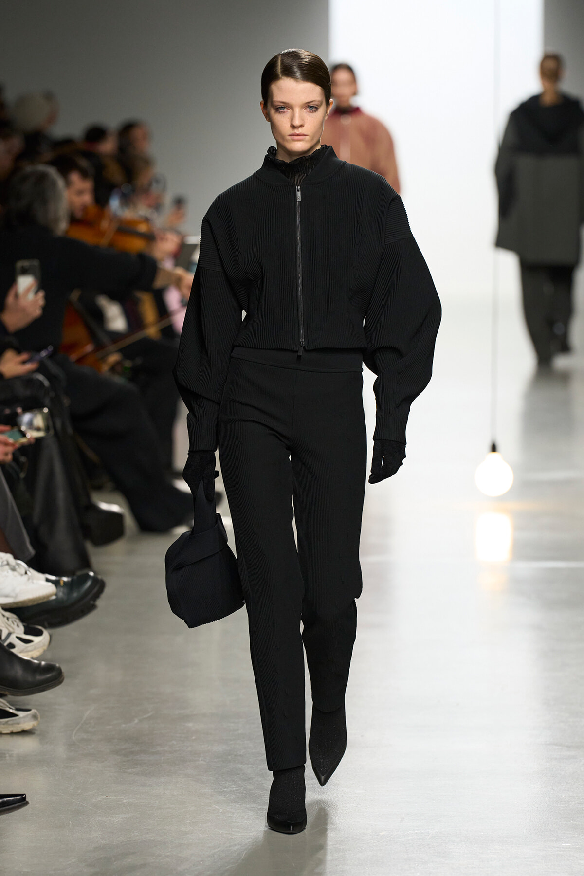 Model walking a runway in an all-black ribbed zip-front top with oversized sleeves and matching pants, holding a black bag.