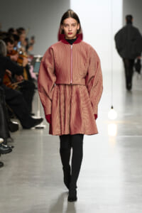 Fashion model walks the runway in a red striped coat with oversized sleeves and burgundy collar, paired with black tights and heeled boots.