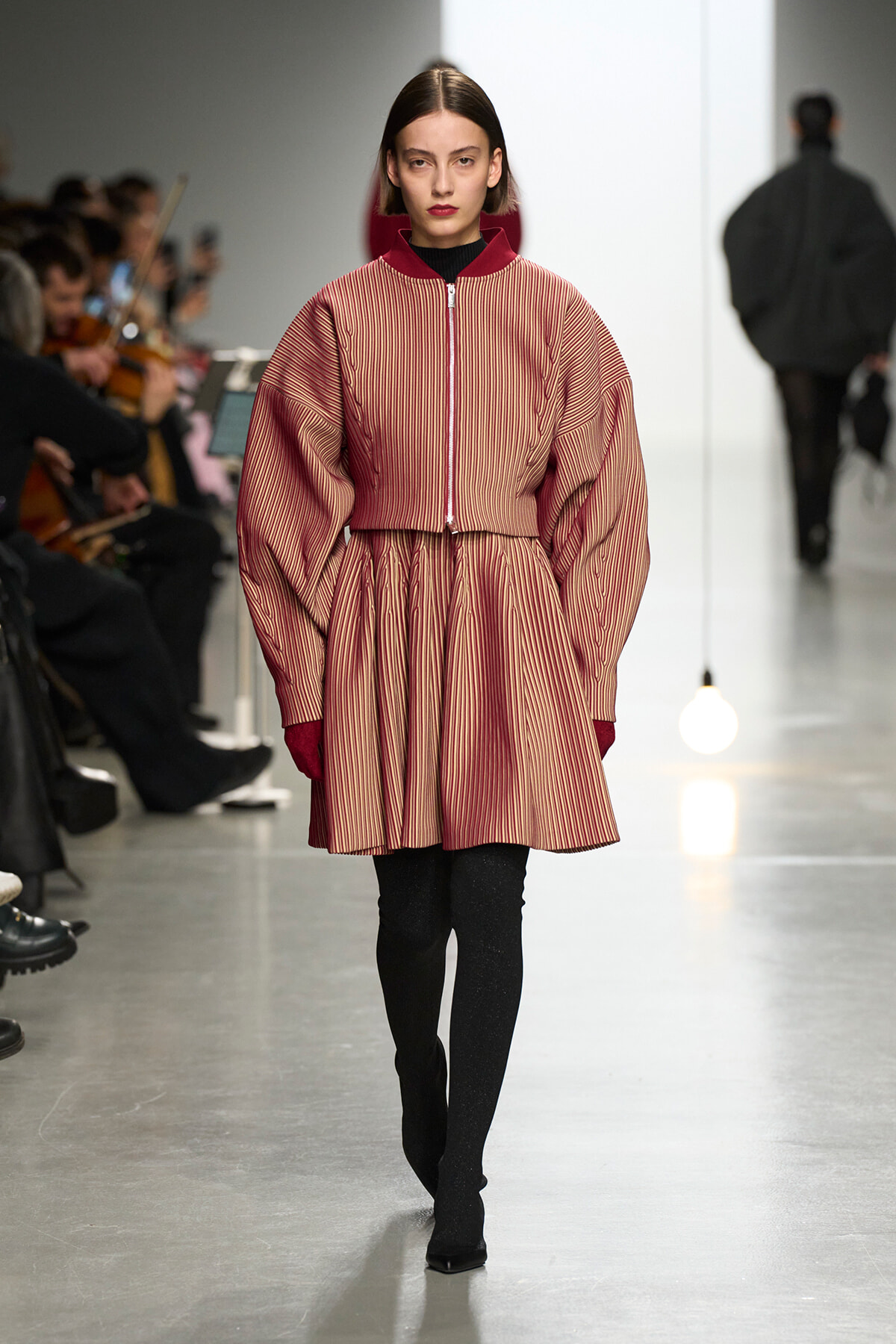 Fashion model walks the runway in a red striped coat with oversized sleeves and burgundy collar, paired with black tights and heeled boots.