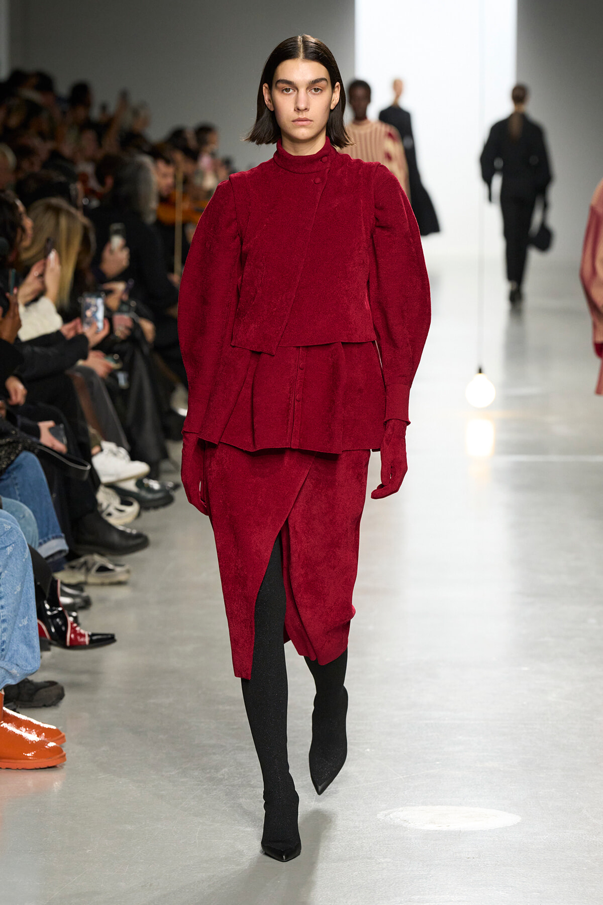 Model on a fashion show runway wearing a burgundy layered velvet top and matching skirt with black tights.