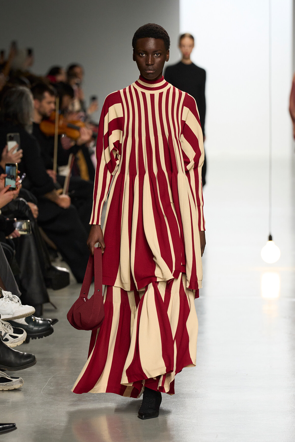 Model on a runway wearing a long red and cream striped knit dress, carrying a matching red bag.