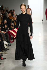 Model walks the runway in a black, long-sleeve knit dress while holding a red-and-cream striped bag; audience watches from the left.