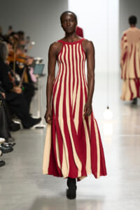 Model walks the runway in a sleeveless red and cream vertical striped maxi dress with a flared hem.