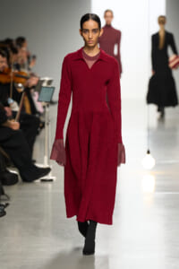Model walking a fashion show in a burgundy knit dress with sheer cuffs, audience on the left and other models in the background.