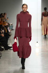 Model walking the runway in a burgundy ribbed turtleneck with a peplum over a matching ribbed skirt and red gloves.