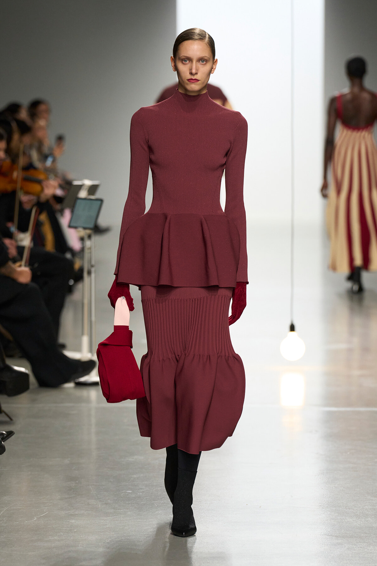 Model walking the runway in a burgundy ribbed turtleneck with a peplum over a matching ribbed skirt and red gloves.