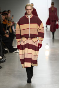 Model walks the runway in a color-block striped burgundy, beige, and blush outfit with a zip-front top and layered skirt; audience visible to the left.