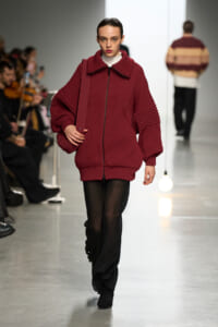 Model walks a runway in a burgundy oversized knit zip jacket with a white turtleneck, carrying a matching shoulder bag.