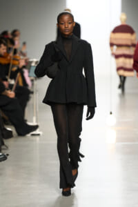 Model walking a fashion runway in a black textured coat and matching pants, wearing gloves and a sheer black turtleneck. Audience seated along the left in a bright studio setting.