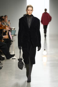 Model walking a fashion show in a black velvet coat over a glittery turtleneck, carrying a textured drawstring bag.